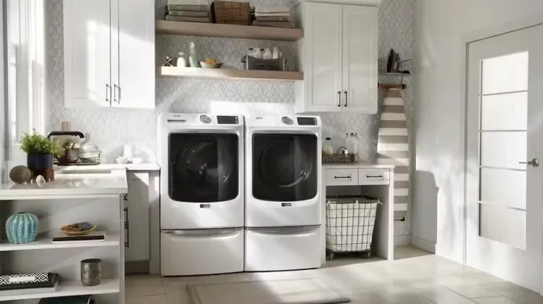Maytag MHW5630HW front-load washer and matching dryer in white, displayed on pedestals in a bright, organized white laundry room